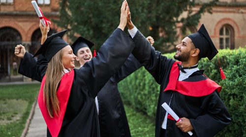 Three happy international graduate friends greeting in university campus in graduation robes with diploma.
